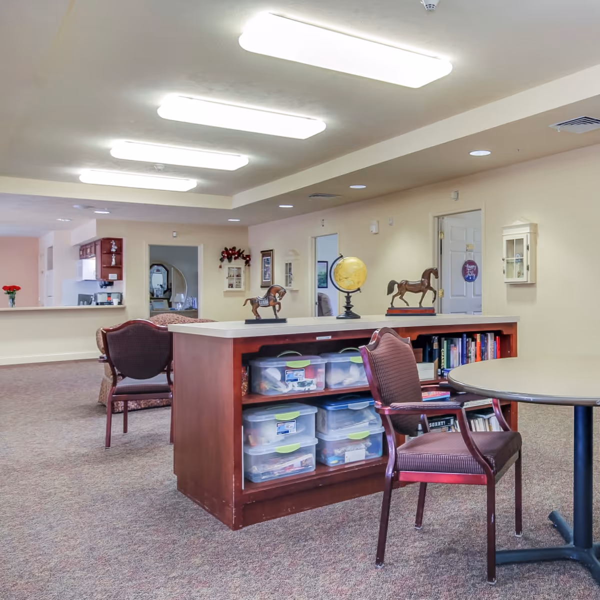 Well-lit assisted living common room with a central shelving island holding plastic bins, books and decorative horse figurines, surrounded by chairs and tables.