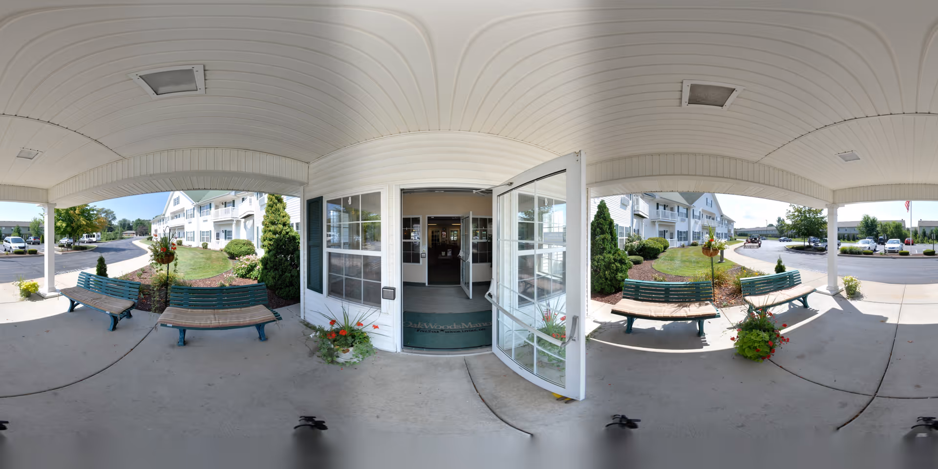 Covered main entrance of a senior living facility with open glass doors, benches, planters, and the building facade and parking lot beyond.