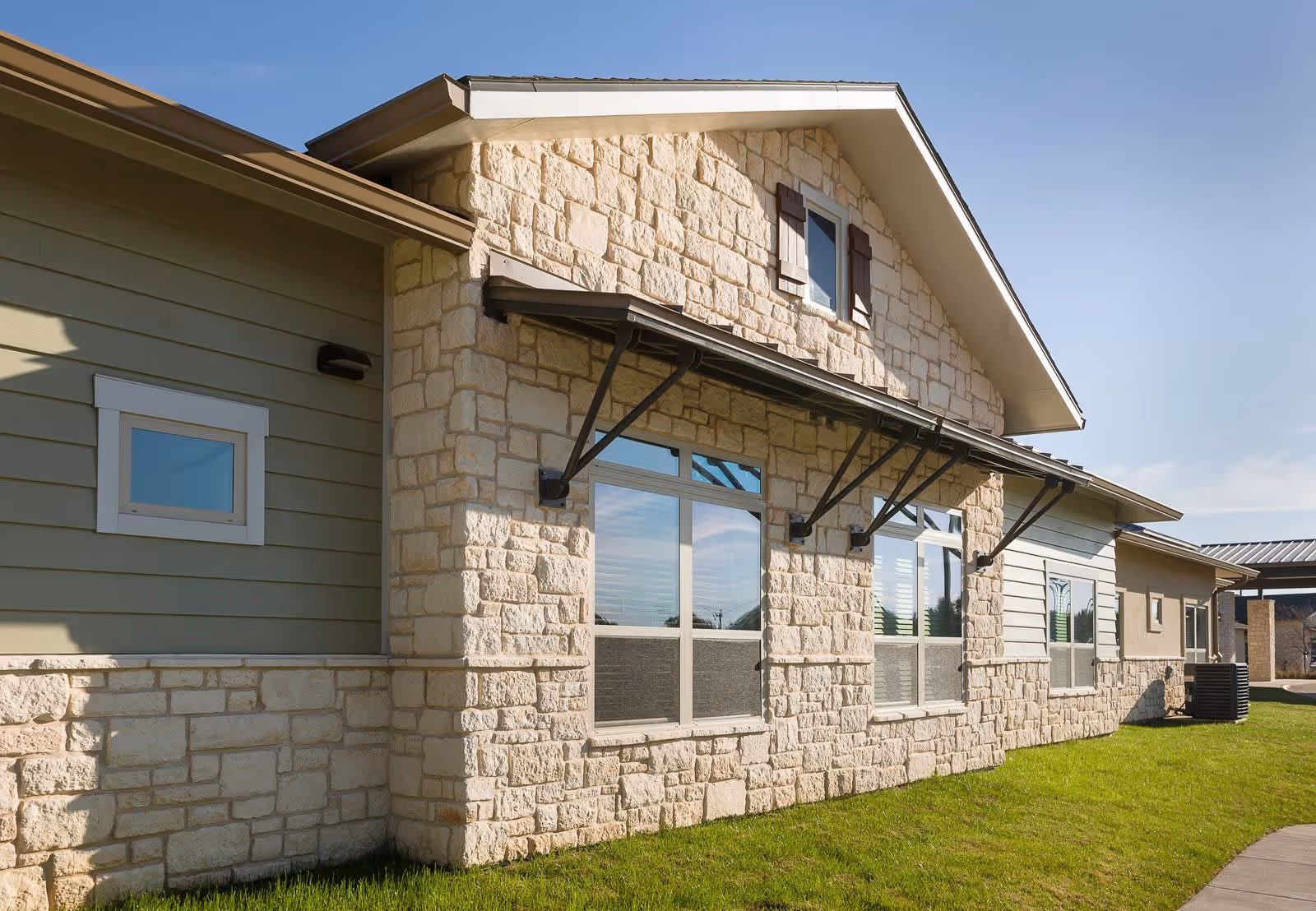 Exterior view of a building with stone and siding walls, multiple windows with awnings, and a well-maintained grassy area in front under a clear blue sky.