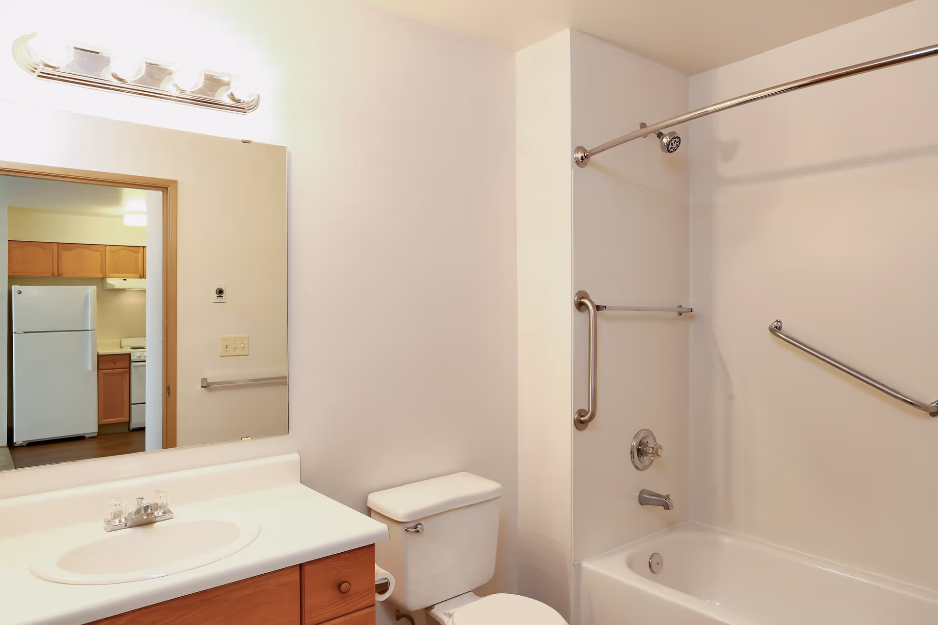 A clean bathroom with a white sink and countertop, wooden cabinets below, a toilet, and a bathtub with a showerhead and grab bars. A large mirror above the sink reflects part of a kitchen with wooden cabinets and a white refrigerator.