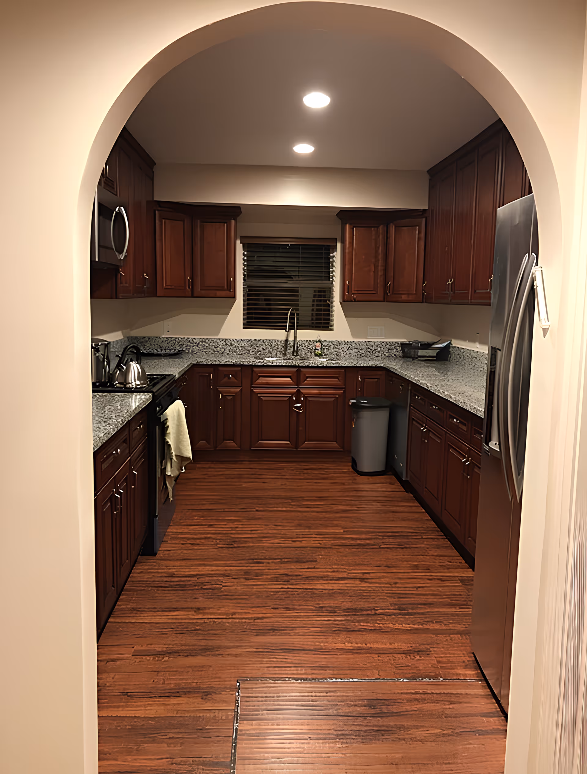 View through an arched doorway into a well-lit kitchen with dark wood cabinets, granite countertops, stainless steel appliances, and wood-look floors.