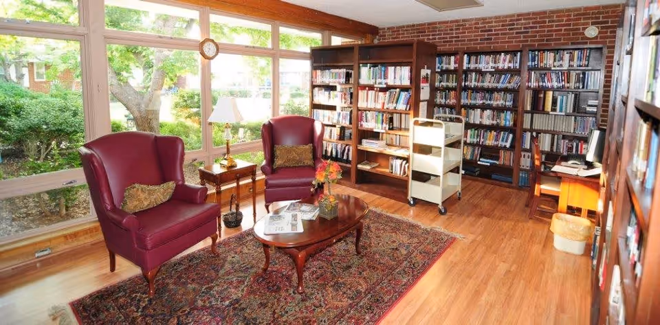 A cozy library room with large windows overlooking greenery outside. The room features two burgundy leather armchairs with decorative pillows, a wooden coffee table with magazines and a flower arrangement, a patterned area rug, wooden bookshelves filled with books, a small side table with a lamp, and a rolling book cart. The floor is wooden, and there is a computer workstation in the corner.