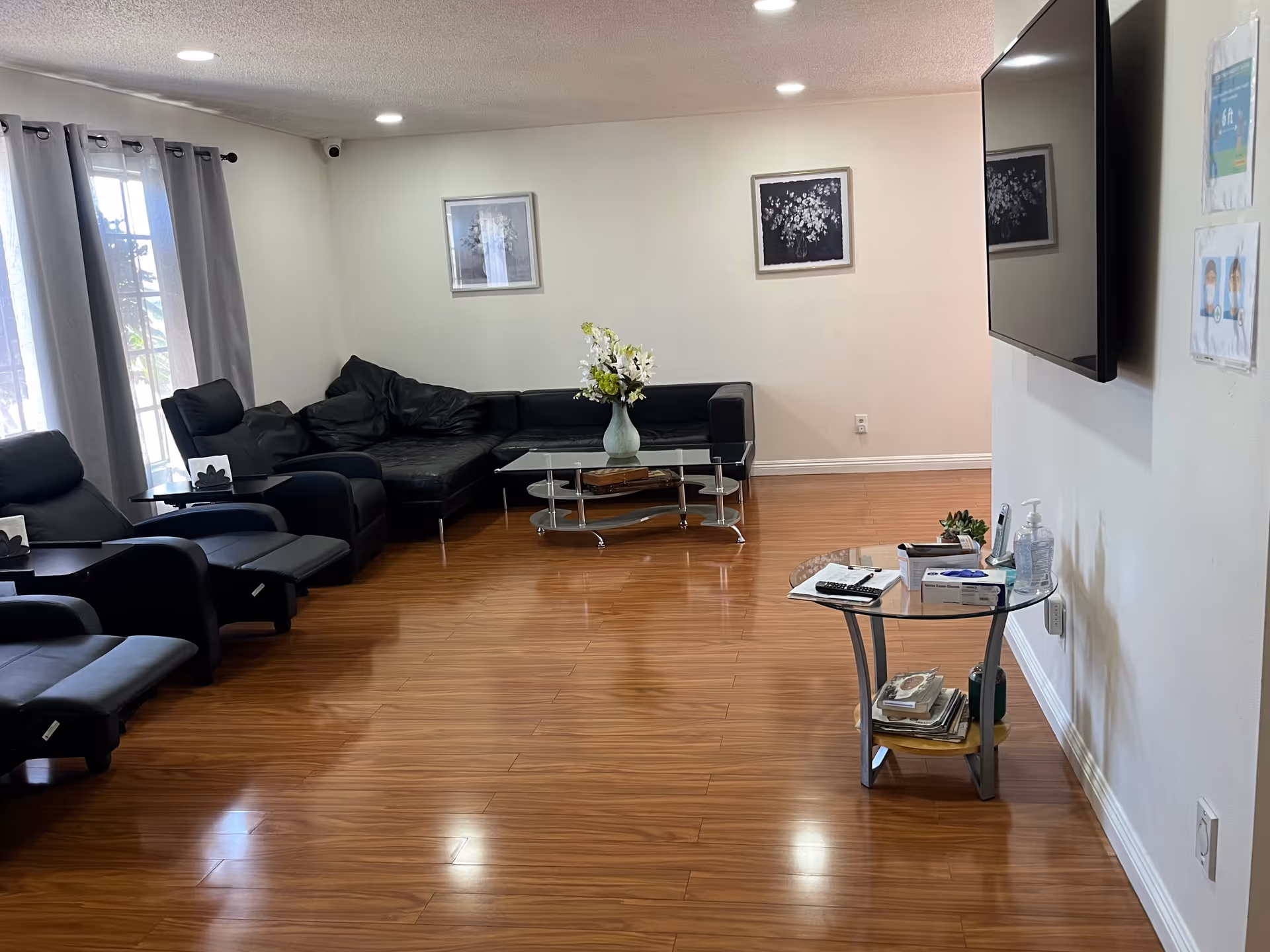 A living room with wooden flooring, black leather sectional sofa and recliners, a glass coffee table with a vase of flowers, a glass side table with various items including a remote control and hand sanitizer, a wall-mounted flat screen TV, two framed floral artworks on the wall, and a window with gray curtains.