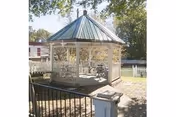 A white wooden gazebo with a green metal roof sits in a fenced yard surrounded by trees.