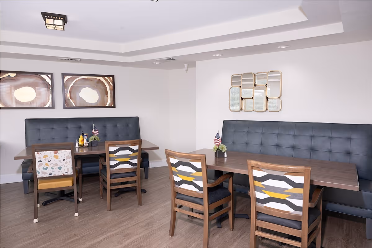 A dining area with two wooden tables, each accompanied by a long black cushioned bench and three wooden chairs with patterned upholstery. The room has light-colored walls decorated with two framed abstract art pieces and a multi-panel mirror. Small American flags and condiment bottles are placed on the tables.