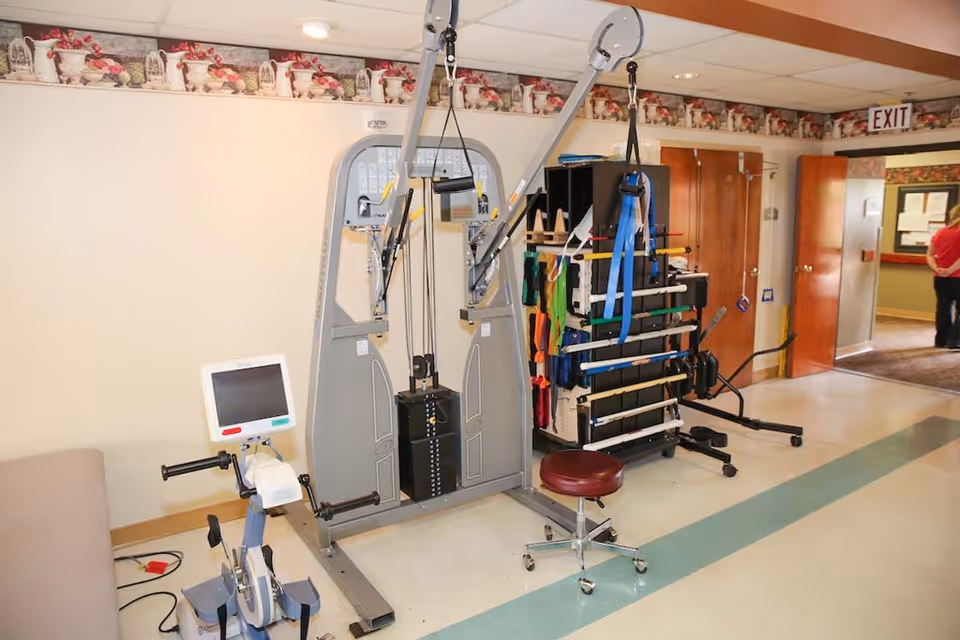 A physical therapy or rehabilitation room in a healthcare facility with exercise equipment including a stationary bike, a multi-function pulley machine, and a rack holding various resistance bands and exercise tools. There is a rolling stool and an open door leading to a hallway with a person standing in the background.