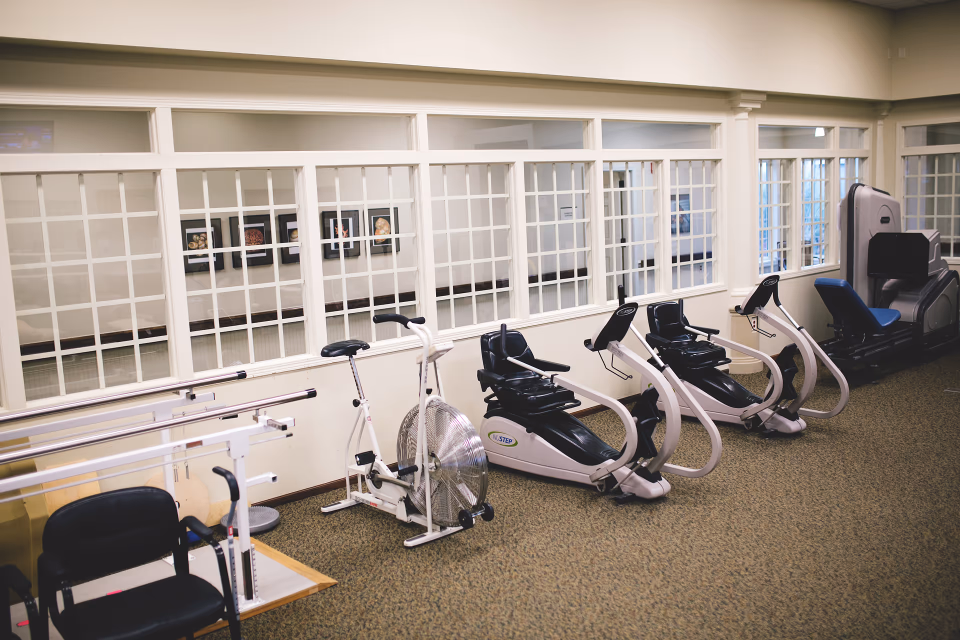 A fitness room with exercise equipment including stationary bikes, recumbent bikes, and a large exercise machine. The room has beige walls, carpeted floor, and large windows with white grid frames looking into another interior space.