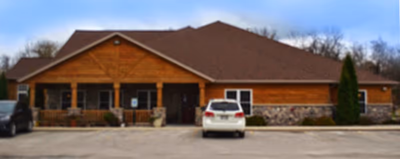 Front exterior view of a single-story senior living facility building with a brown roof, wooden siding, stone accents, a covered porch, and a parking lot with two cars parked in front.