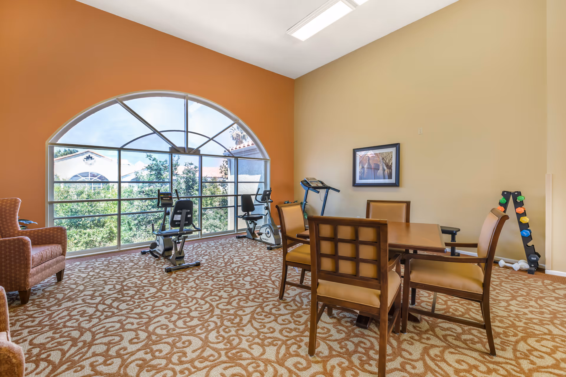 Bright multipurpose common room with a table and chairs, exercise equipment, and a large arched window.