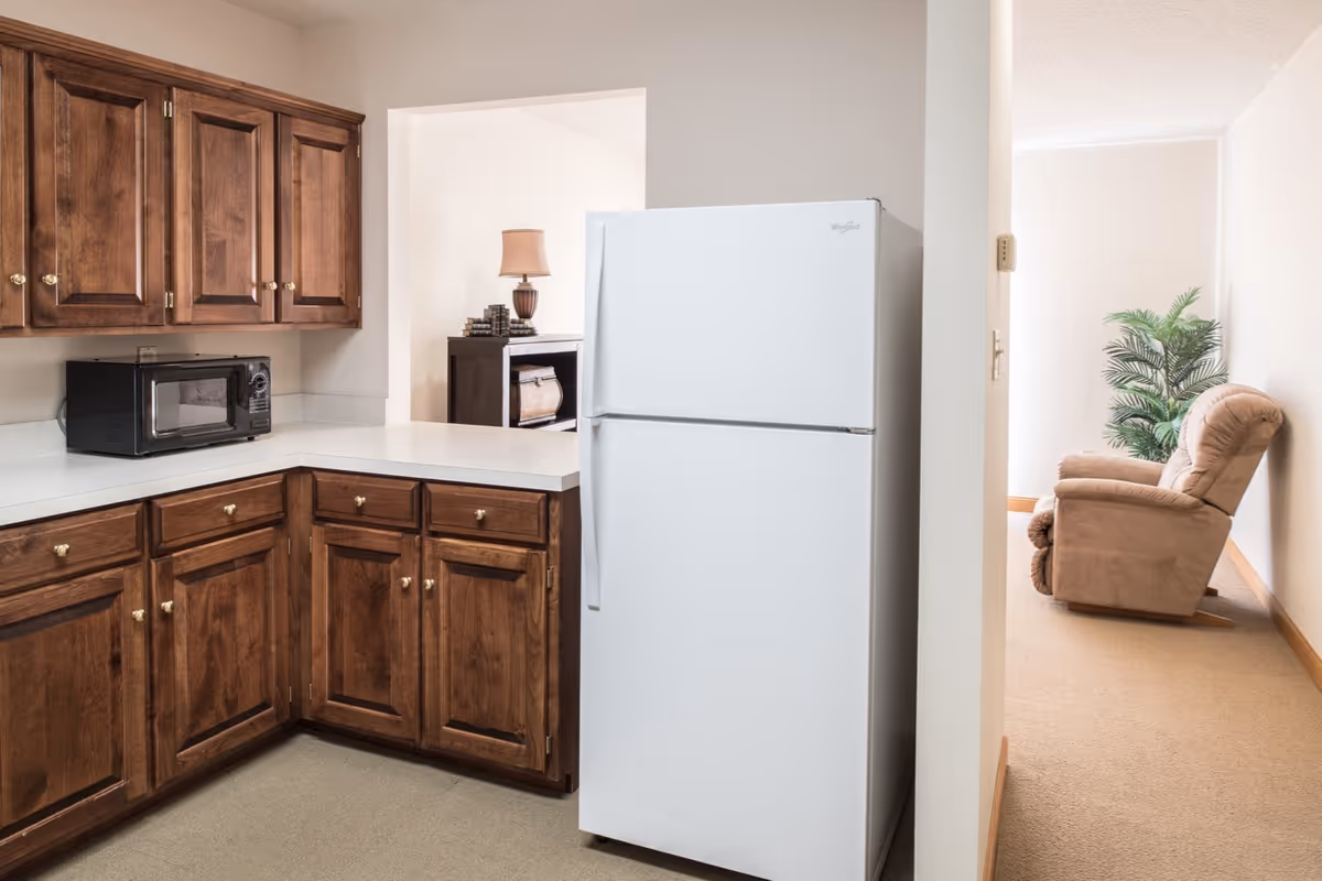 Interior view of a senior living facility kitchen with wooden cabinets, a white refrigerator, and a black microwave on the countertop. Adjacent to the kitchen is a hallway leading to a room with a beige recliner chair and a green potted plant.