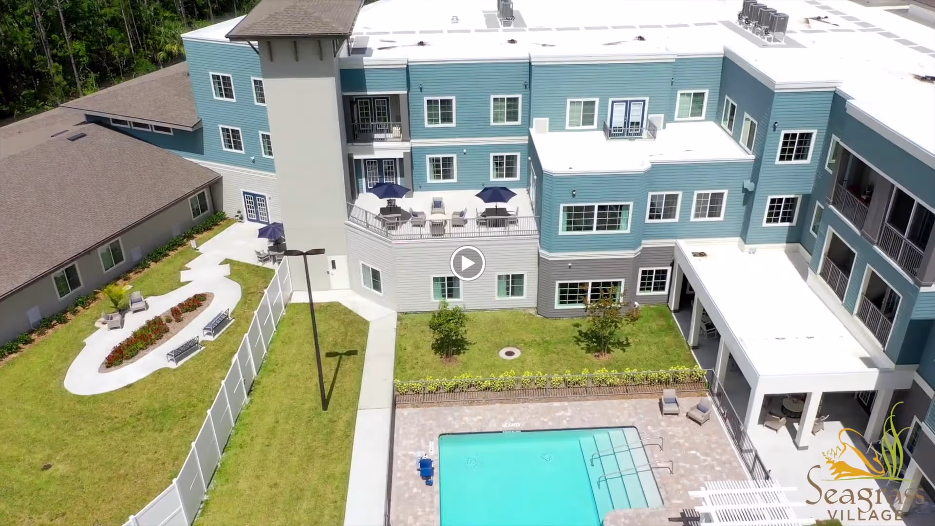 Aerial view of Seagrass Village of Port Orange showing a multi-story building with blue and gray exterior walls, a fenced garden area with benches and plants, a swimming pool with lounge chairs, and a patio with tables and umbrellas.