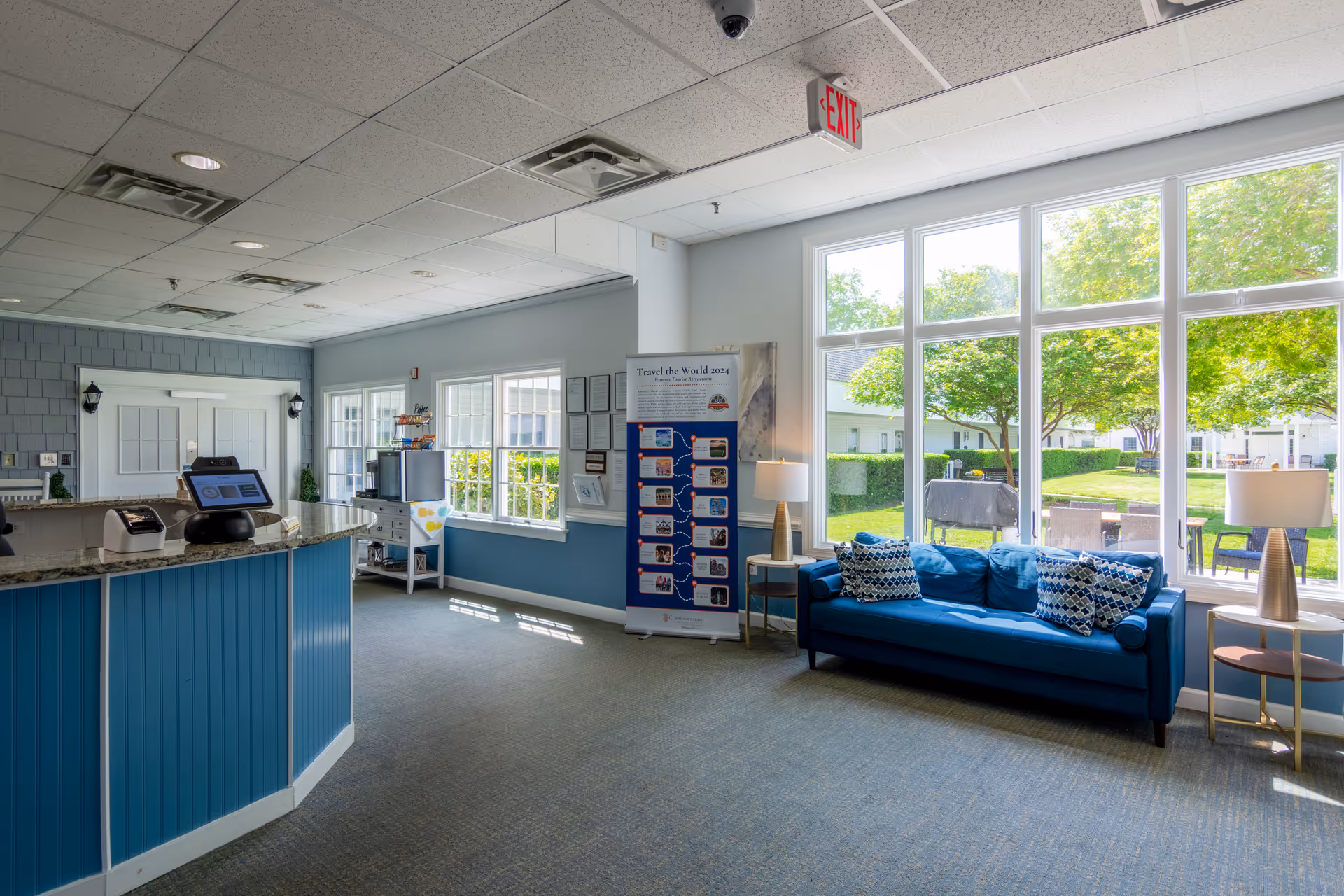 Bright reception and sitting area with a blue front desk and matching blue sofa by large windows overlooking a garden.