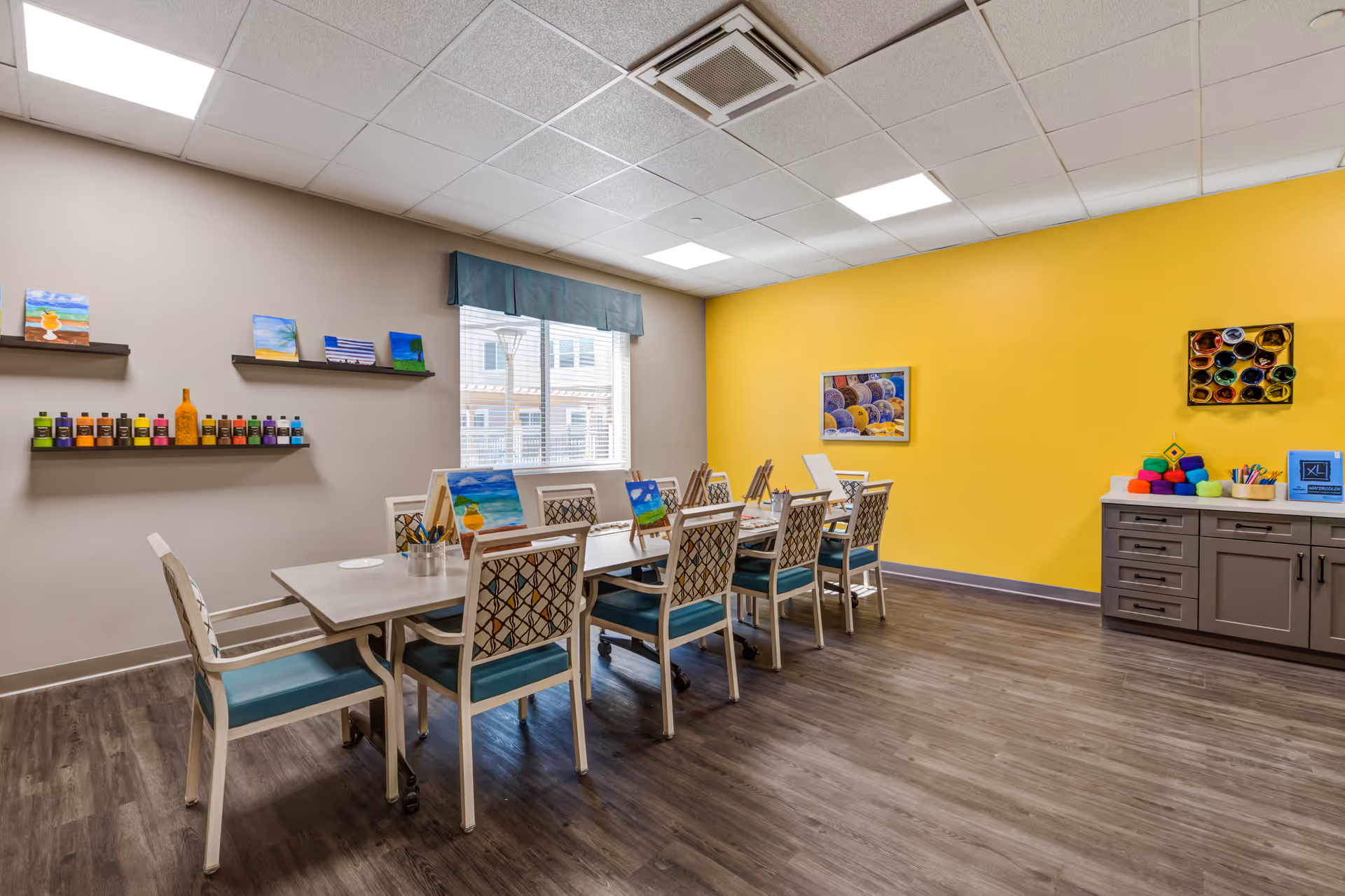 Bright activity room with a long table and chairs set up for painting, shelves of paint bottles, and a yellow accent wall.