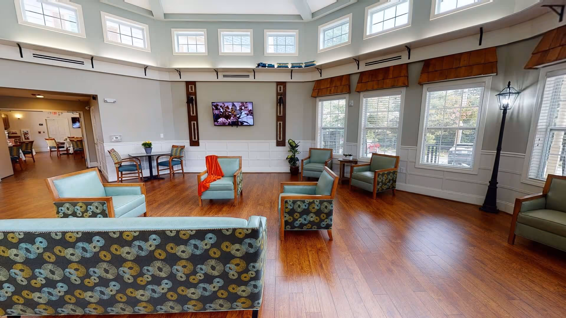 A spacious and well-lit common area with wooden flooring, multiple armchairs and sofas with patterned upholstery, a small table with two chairs, large windows with blinds, a wall-mounted TV displaying a nature scene, and a tall decorative lamp post inside the room.