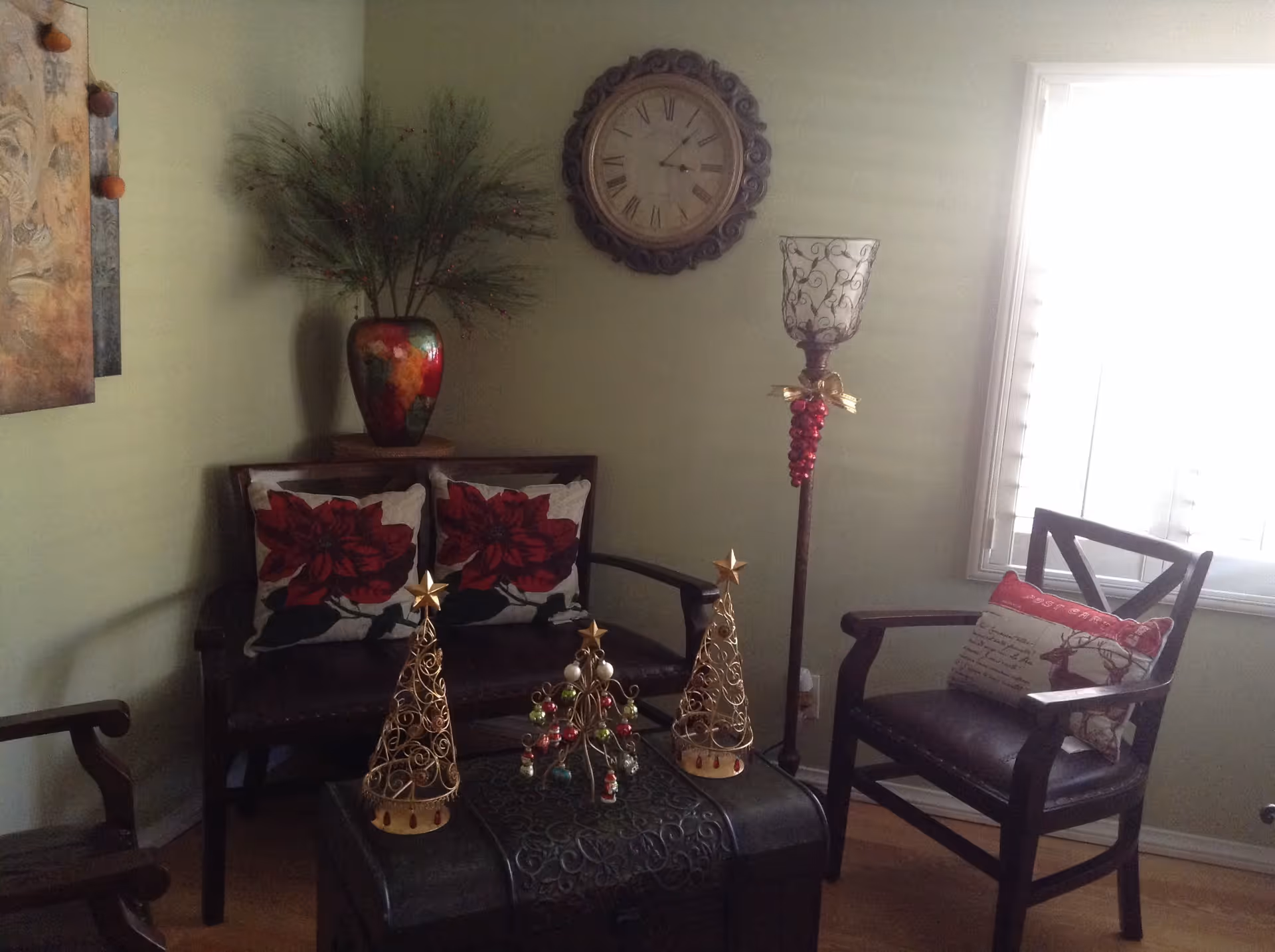 A cozy living room corner with a dark wooden loveseat adorned with two red floral pillows, a matching wooden armchair with a festive pillow, a decorative clock on the wall, a tall candle holder decorated with red berries, and a table with three small decorative Christmas trees. A colorful vase with greenery is placed on a small table behind the loveseat.