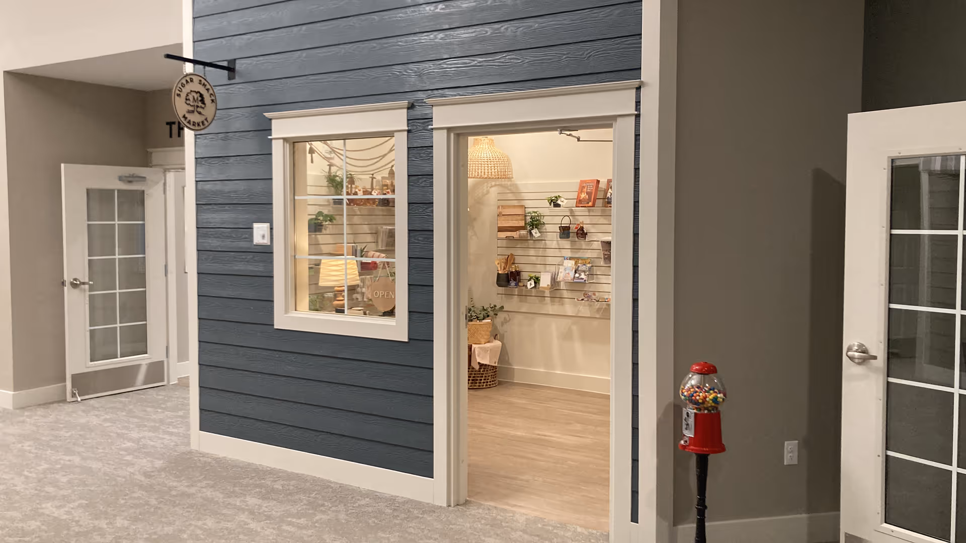 Interior view of a room designed to look like a small storefront with blue siding, white trim, a window, and an open doorway showing shelves with various items inside. There is a gumball machine to the right and a door with glass panes on the left side of the image.