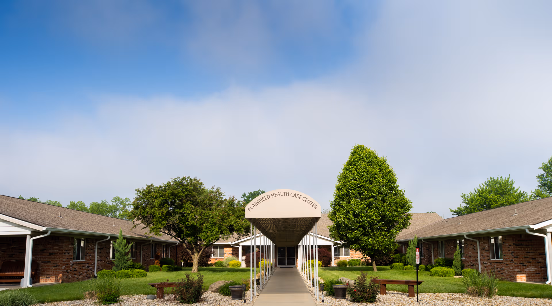 Front entrance of Plainfield Health Care Center with a covered walkway between low brick buildings and landscaped grounds.