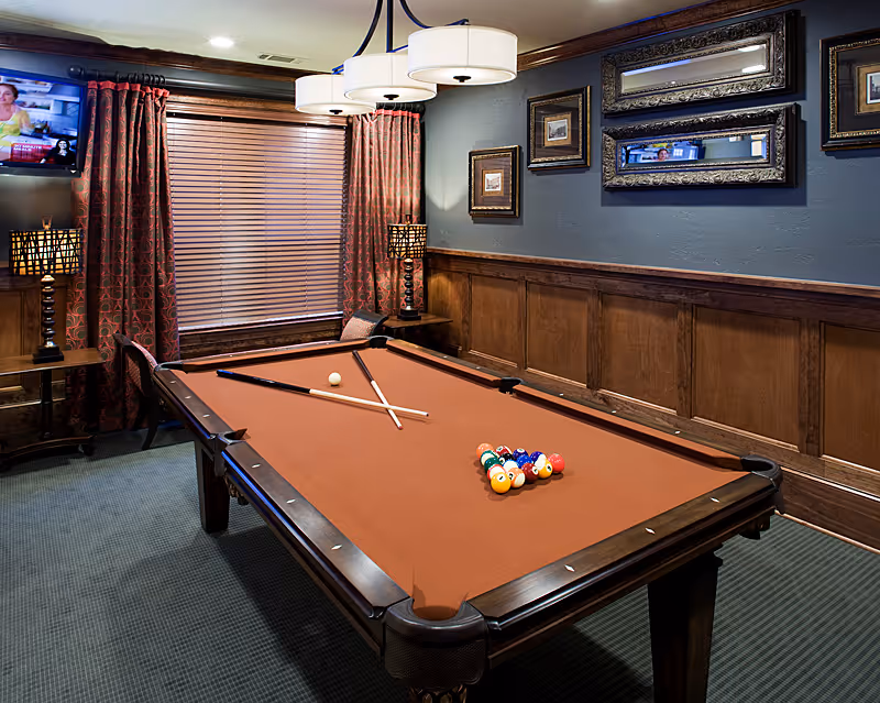 A cozy game room with a brown felt pool table in the center, two pool cues and a white cue ball on the table, a rack of colorful billiard balls arranged for a game. The room has wood-paneled walls with framed pictures and a large mirror, a window with closed blinds and red patterned curtains, two table lamps with patterned shades on side tables, a wall-mounted TV in the corner, and a modern ceiling light fixture above the pool table.