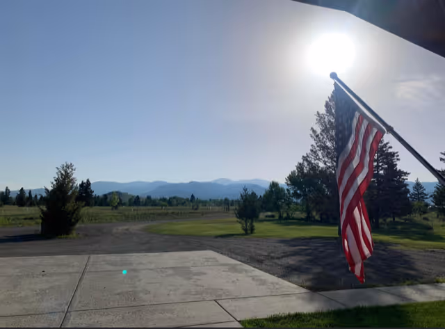 American flag on a porch pole beside a concrete walkway and lawn, with a gravel driveway, trees and distant mountains under a bright sun.