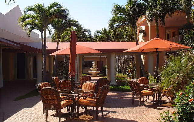 Outdoor courtyard with wicker patio tables and chairs under red umbrellas, palm trees, and a central fountain.