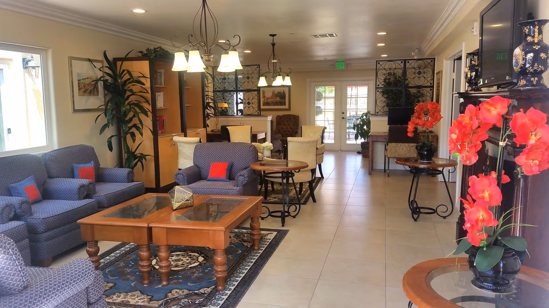 A bright and spacious living room area in an assisted living facility featuring multiple upholstered armchairs and sofas with colorful cushions, wooden coffee tables on a patterned rug, decorative plants including vibrant red flowers, wall art, and a television mounted above a fireplace. The room has tiled flooring, pendant lighting, and large windows allowing natural light to enter.
