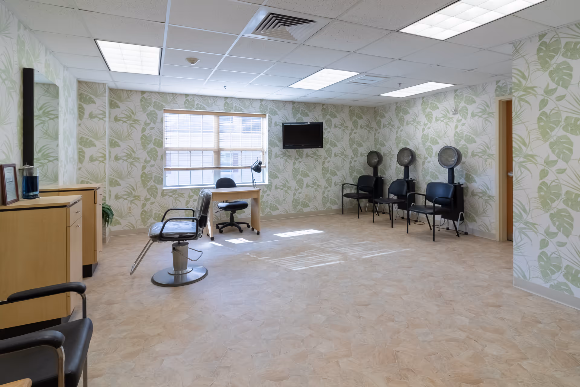 A spacious room with light-colored flooring and walls decorated with green leafy patterned wallpaper. The room contains a salon chair, a desk with an office chair and lamp near a window with blinds, a wall-mounted TV, and three black chairs with hair dryers behind them. There is also a wooden cabinet with a mirror on the left side.
