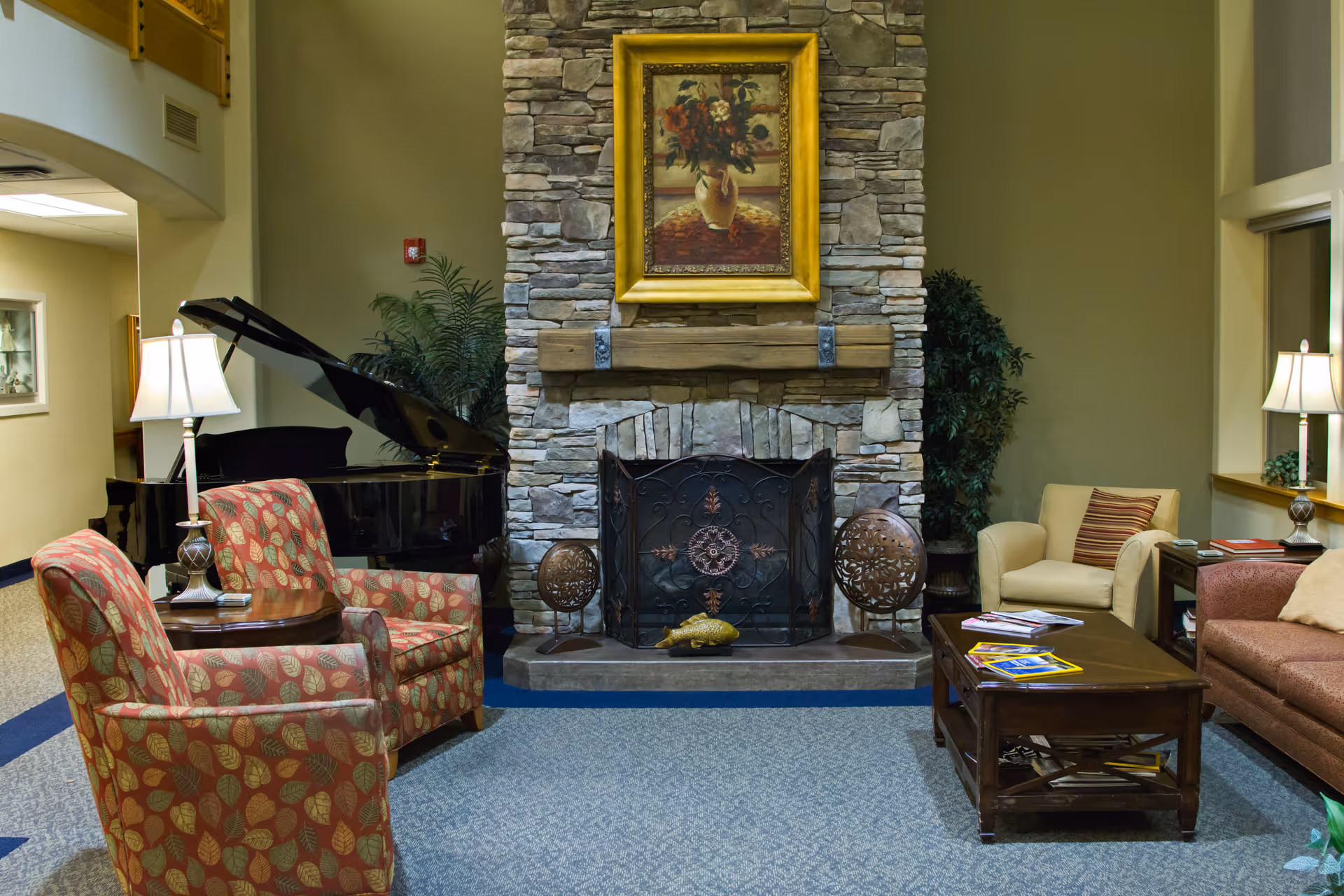 A cozy living room area with a stone fireplace in the center, decorated with a framed painting of flowers above the mantel. Two patterned armchairs with a small round table and lamp are on the left side, next to a black grand piano. On the right side, there is a beige armchair, a brown sofa, a wooden coffee table with magazines, and a side table with a lamp. The room has carpeted flooring and plants near the fireplace.