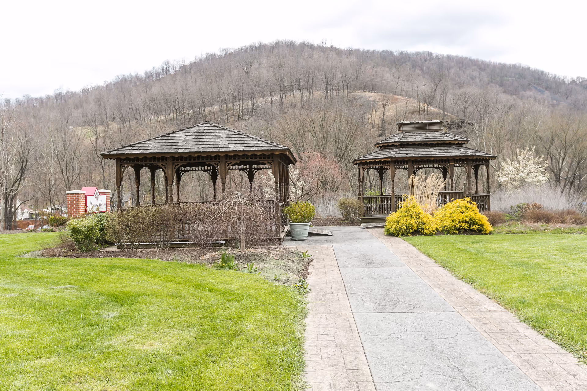 A landscaped outdoor area with two wooden gazebos connected by a paved walkway. The scene includes green grass, shrubs, and trees with a backdrop of a wooded hill under an overcast sky.