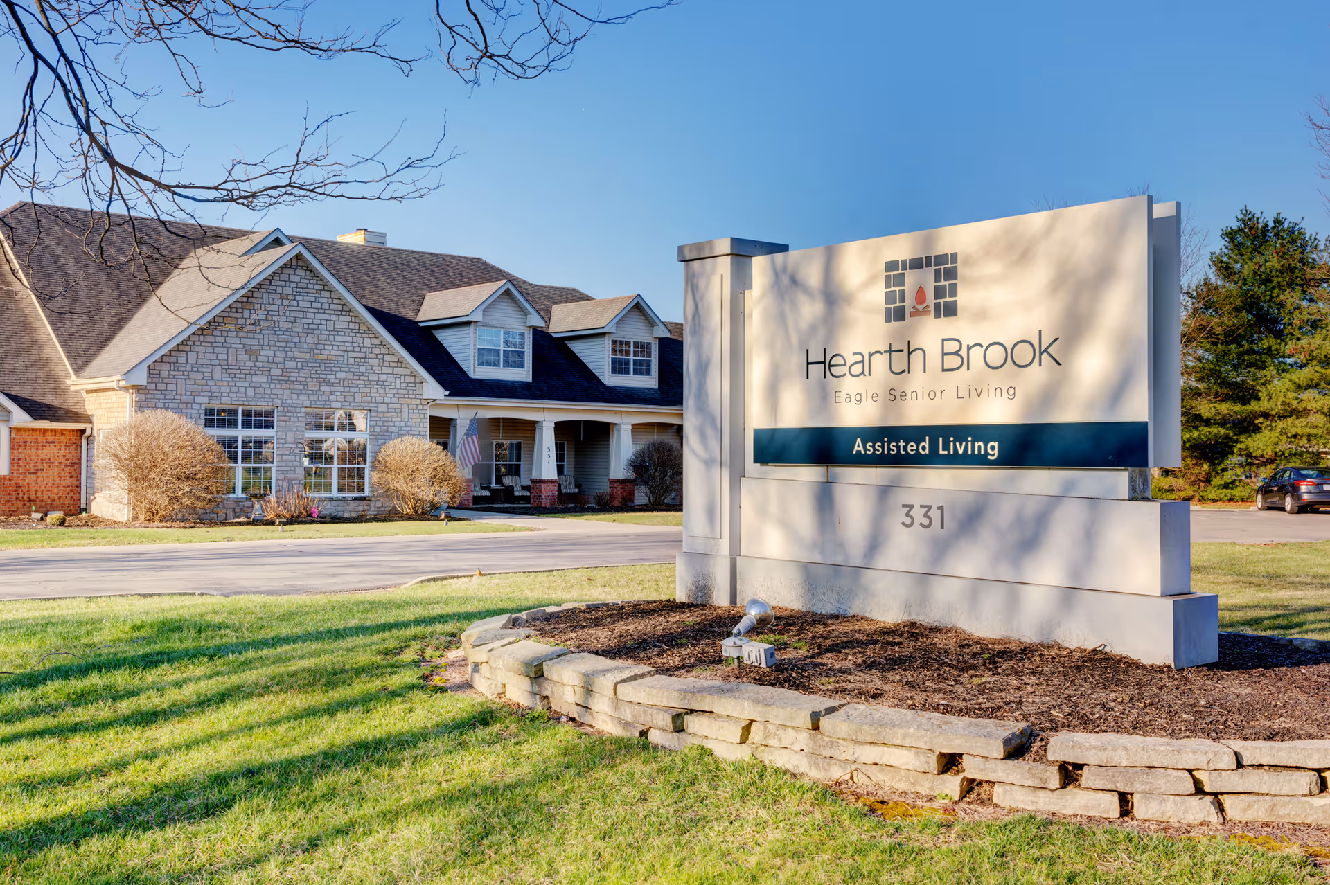 A large stone sign reading 'Hearth Brook Assisted Living' stands on the lawn in front of the Hearth Brook senior living building.