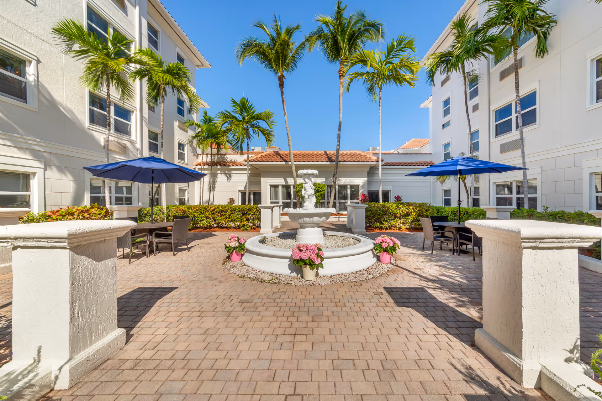 Outdoor courtyard area at Brookdale Deer Creek featuring a central white fountain surrounded by pink flowers, palm trees, and patio tables with blue umbrellas. The courtyard is paved with bricks and flanked by white buildings under a clear blue sky.