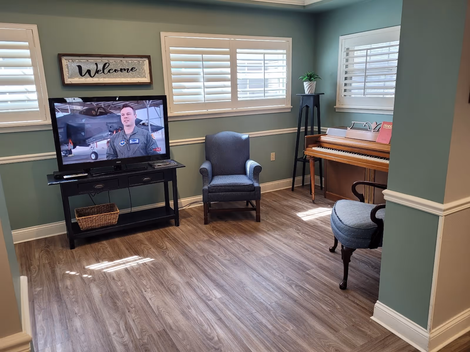 A cozy living room area with teal walls and wood flooring. The room features a flat-screen TV on a black stand displaying a man in a military uniform, a blue upholstered armchair, a wooden piano with a music book on top, a small plant on a tall black stand, and a blue cushioned chair with wooden arms. There are three windows with white plantation shutters and a framed 'Welcome' sign above the TV.