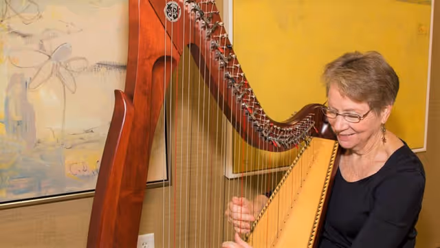 An elderly woman with short hair and glasses is playing a large wooden harp indoors. She is smiling and appears focused on the instrument. Behind her, there are two abstract paintings on a yellow and beige wall.
