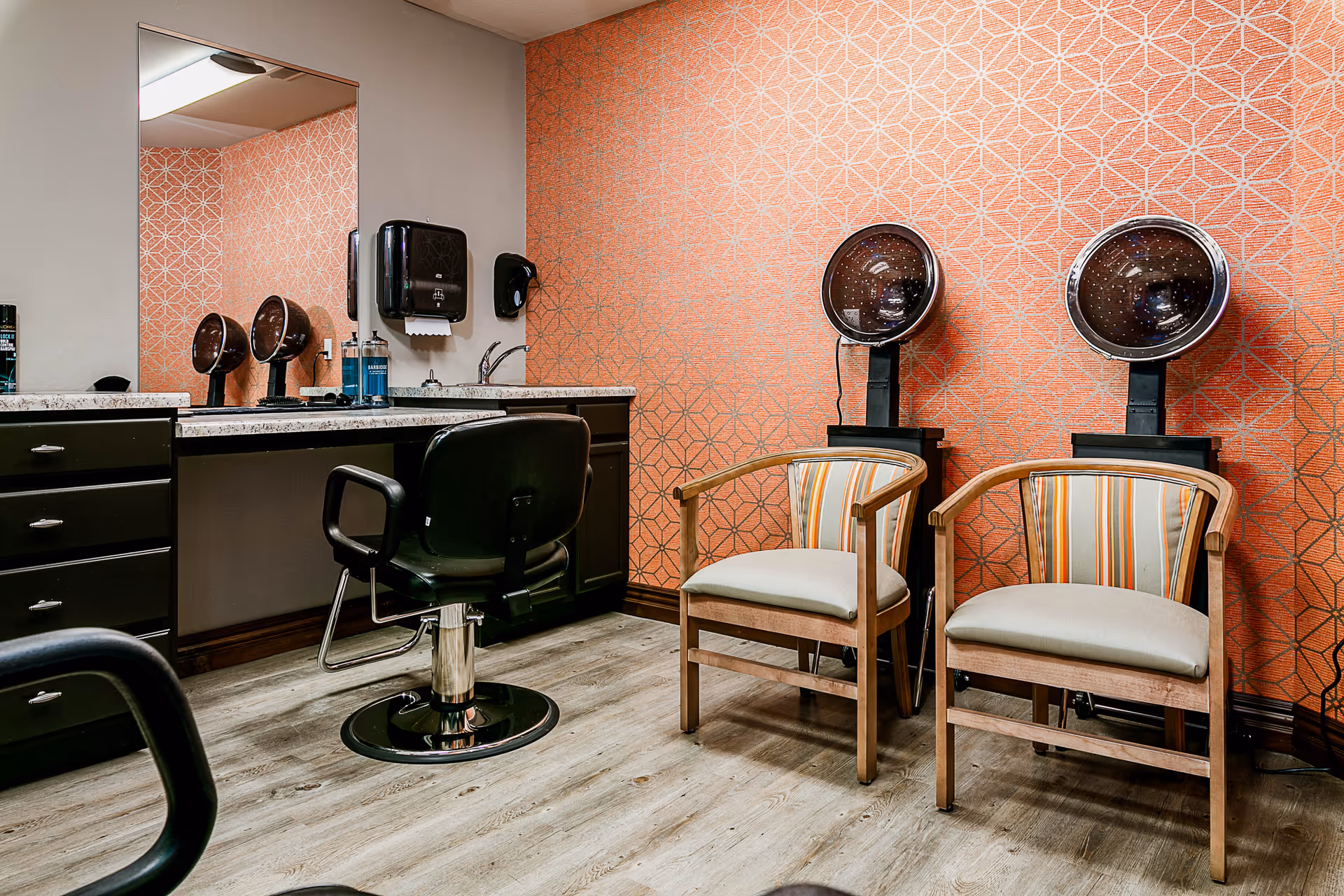 Interior view of a salon area with two black salon chairs in front of a counter with a large mirror, two hair dryers mounted on stands, and two wooden chairs with striped cushions against an orange patterned wall.