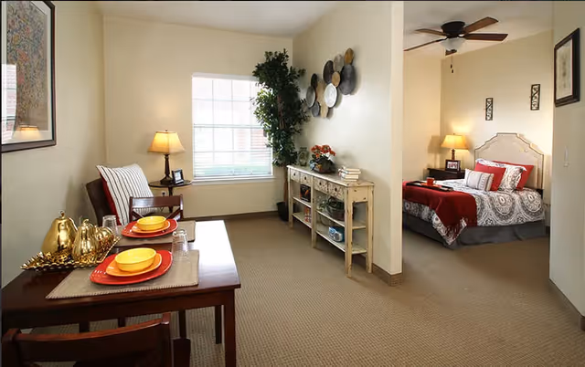 Bright apartment interior showing a dining table set for two, a small seating area, a decorative console, and a bedroom with a bed and lamps.
