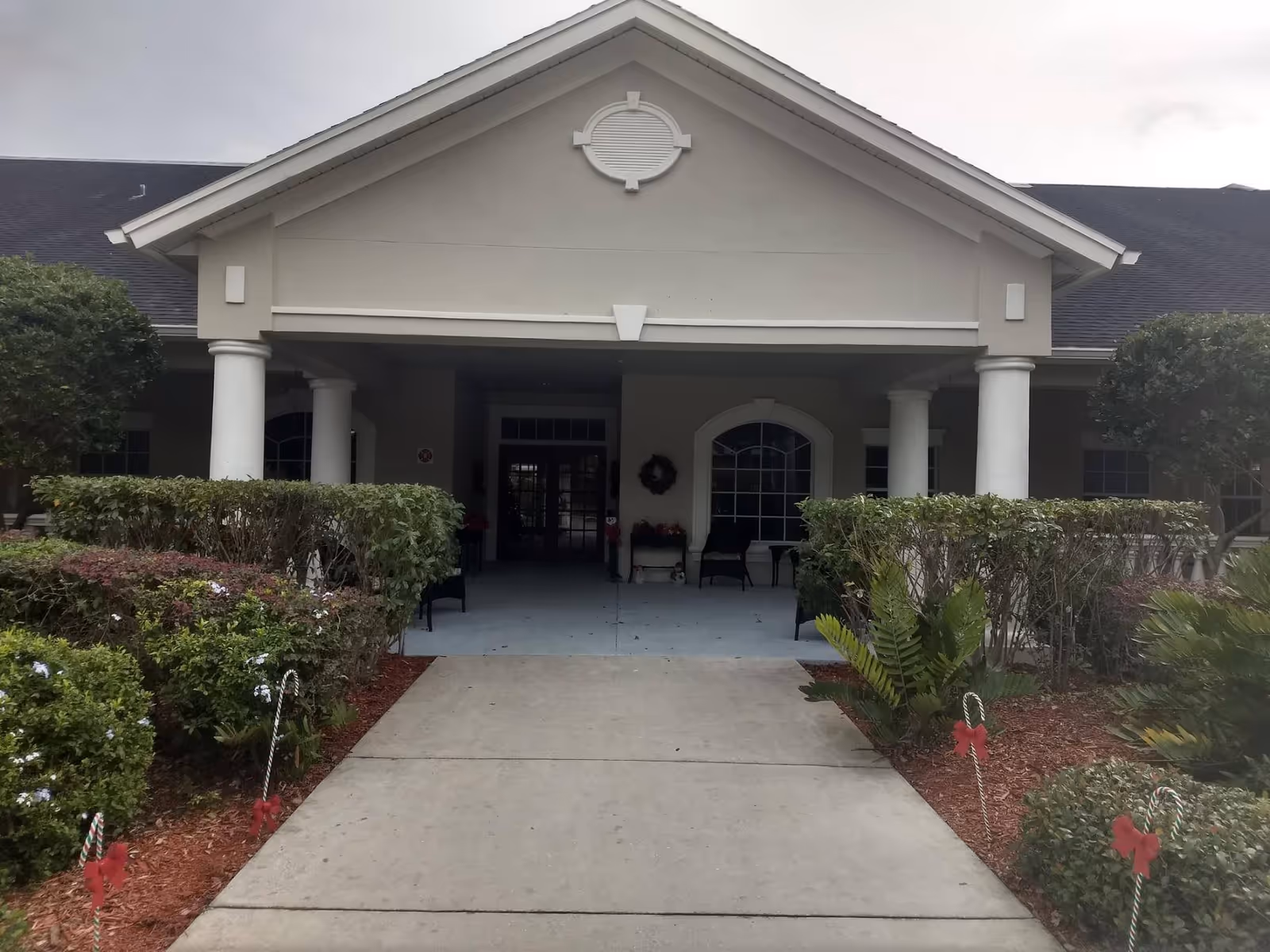 Entrance of a building with a covered porch supported by white columns, surrounded by neatly trimmed bushes and small trees. The walkway leading to the entrance is decorated with candy cane stakes with red bows.