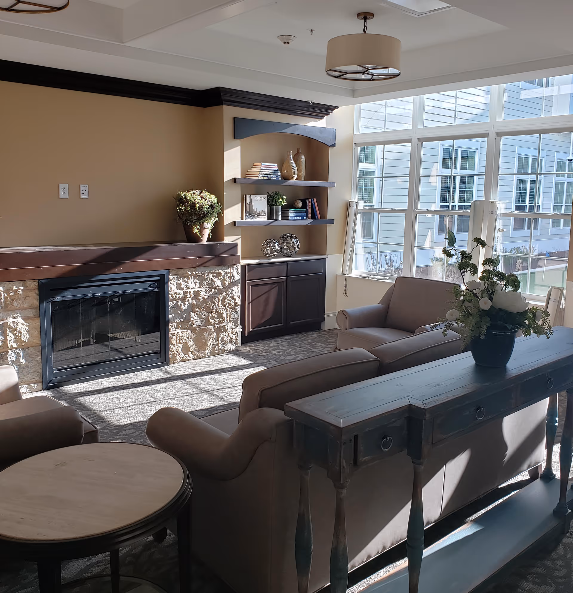 A cozy living room area with beige walls and a stone fireplace. There are two beige armchairs and a beige sofa arranged around a wooden console table with a flower vase on it. Built-in shelves with decorative items and books are on the wall next to large windows letting in natural light.