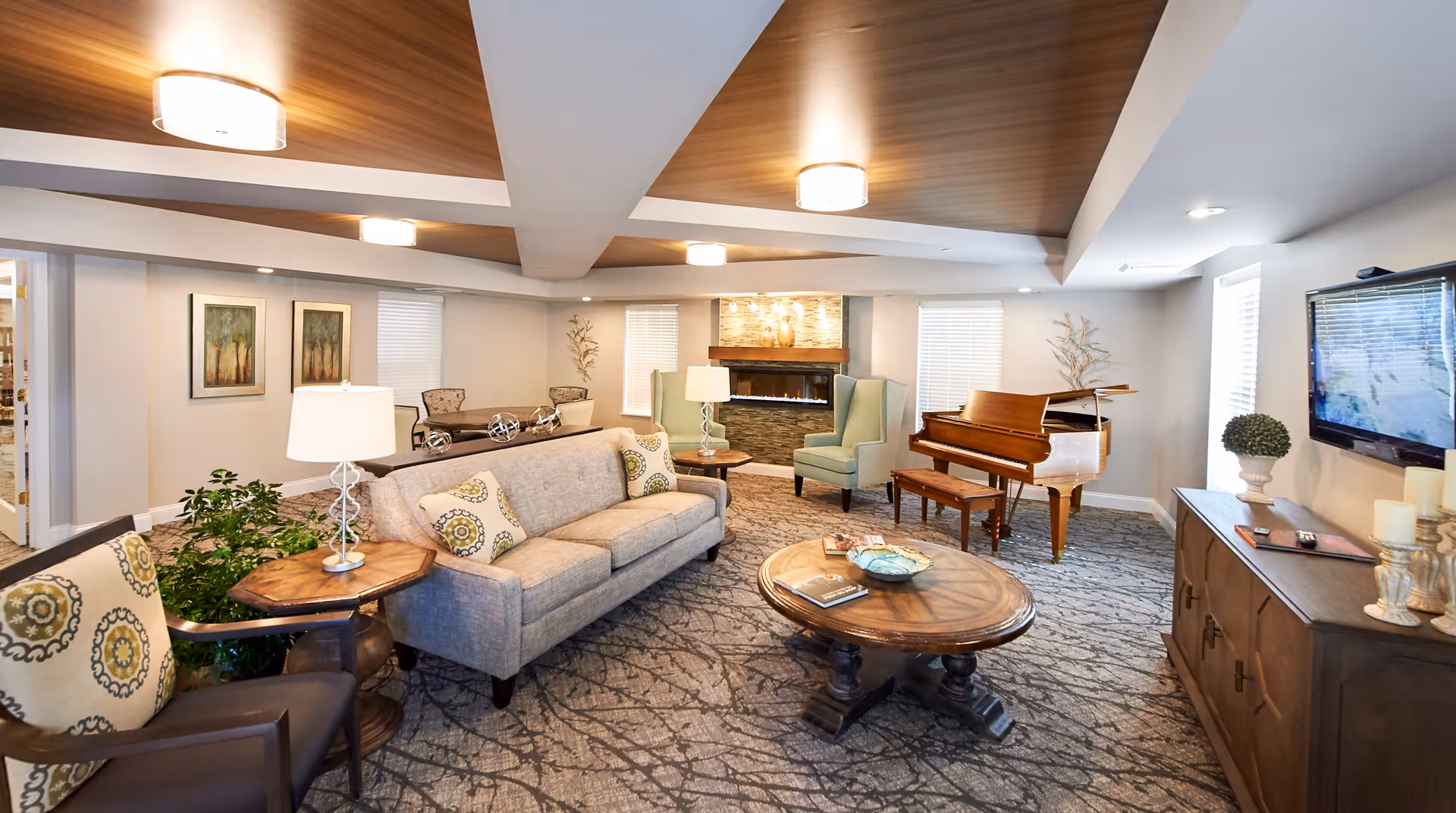 A cozy living room area with a gray sofa, patterned armchairs, wooden coffee and side tables, a flat-screen TV mounted on the wall, a grand piano, and a modern fireplace with stone surround. The ceiling has wood paneling and recessed lighting, and the room is decorated with plants, artwork, and decorative objects.