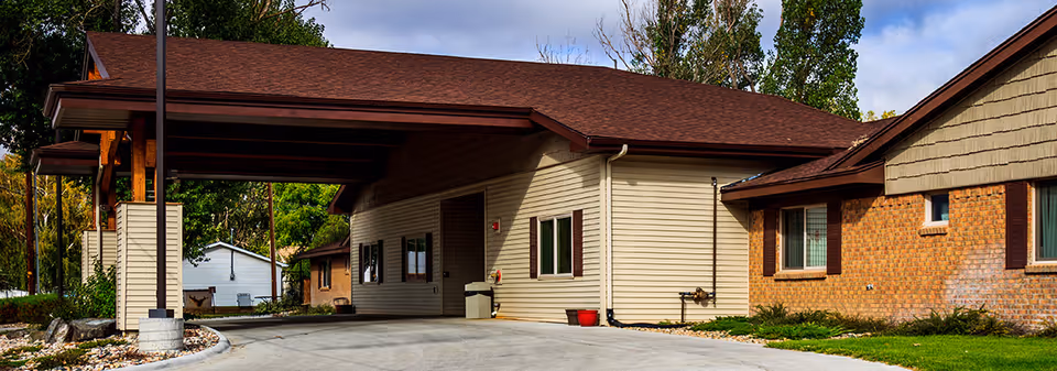 Exterior view of Douglas Care Center showing a covered driveway entrance with a brown roof, beige siding, and brick walls. There are windows with brown shutters and some greenery around the building.