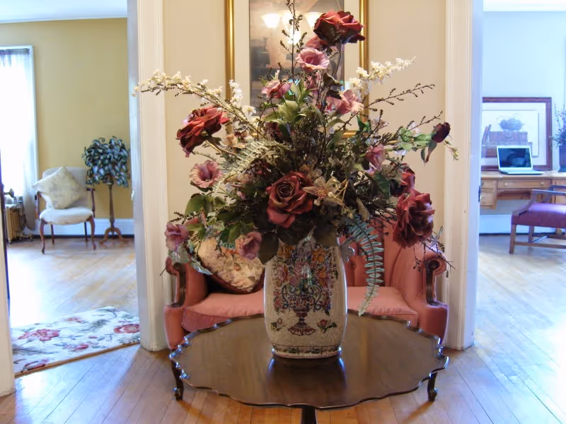 A large floral arrangement with red and pink flowers in a decorative vase sits on a wooden table in the center of a room. Behind the table is a pink upholstered loveseat with a floral pillow. The room has wooden floors and is connected to two adjacent rooms, one with a white armchair and a small plant, and the other with a wooden desk, chair, and a laptop.