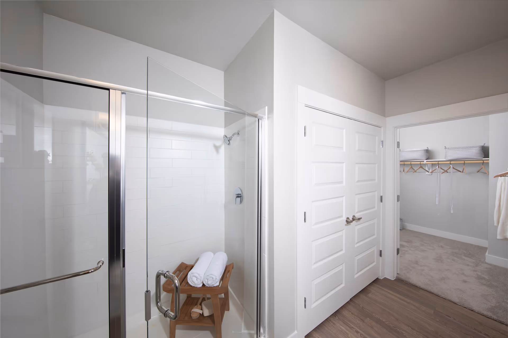 A modern bathroom featuring a glass-enclosed shower with white tiled walls. Inside the shower, there is a wooden bench holding two rolled white towels and a brush. Adjacent to the shower is a white double-door closet, and an open doorway reveals a carpeted walk-in closet with hanging rods and storage baskets.
