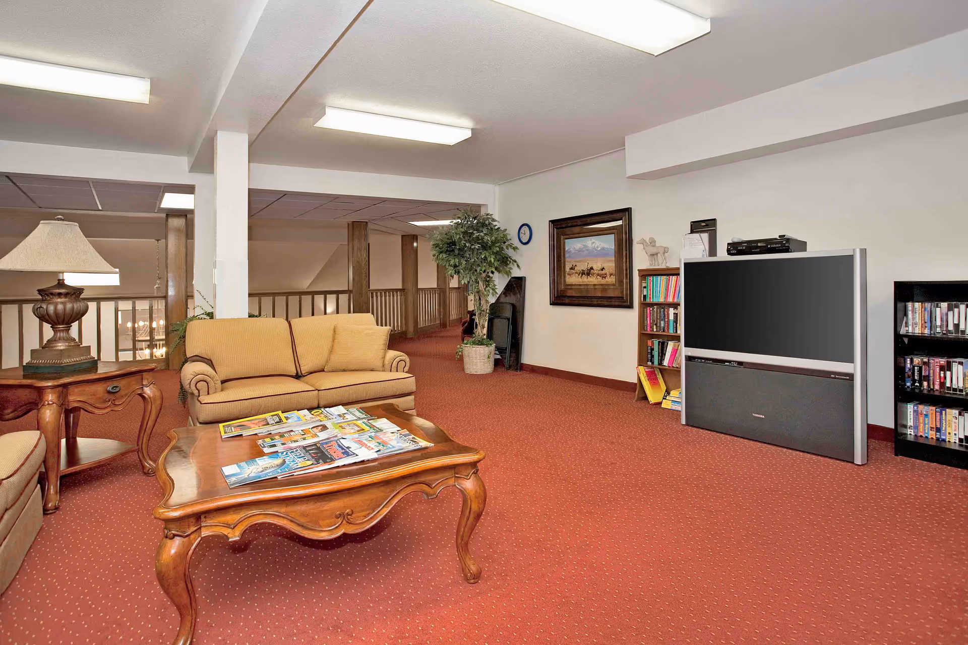Communal living room area with sofas, a wooden coffee table with magazines, a large TV and bookshelves on red carpet.