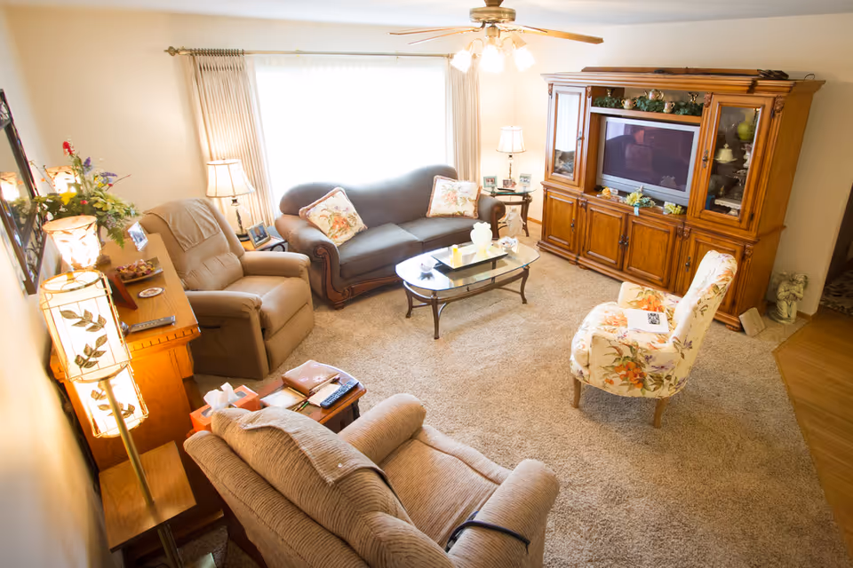 Well-lit cozy living room with a sofa, recliners, a floral armchair, glass coffee table, and a large wooden entertainment center.