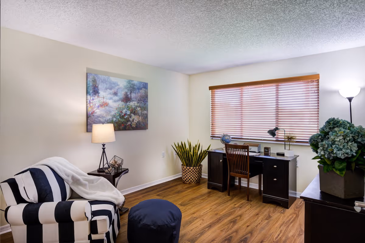 A cozy room with a black and white striped armchair draped with a white blanket, a small side table with a lamp and decorative items, a dark wooden desk with a chair positioned in front of a window with wooden blinds, a potted plant on the floor, and a floor lamp next to a cabinet with a large potted plant on top. A colorful landscape painting hangs on the wall above the armchair.