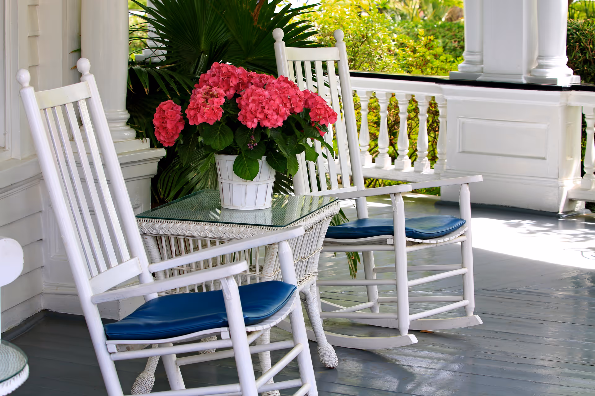A porch area with two white wooden rocking chairs with blue cushions, a white wicker table with a glass top, and a white pot of vibrant pink flowers. The porch has white railings and columns, with greenery visible in the background.