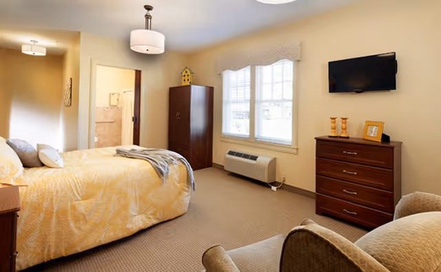 A cozy bedroom with a bed covered in yellow patterned bedding, a wooden wardrobe, a chest of drawers with decorative items, a wall-mounted TV, a window with blinds, and an armchair. The room has beige walls and carpeted floor, with a doorway leading to a bathroom.