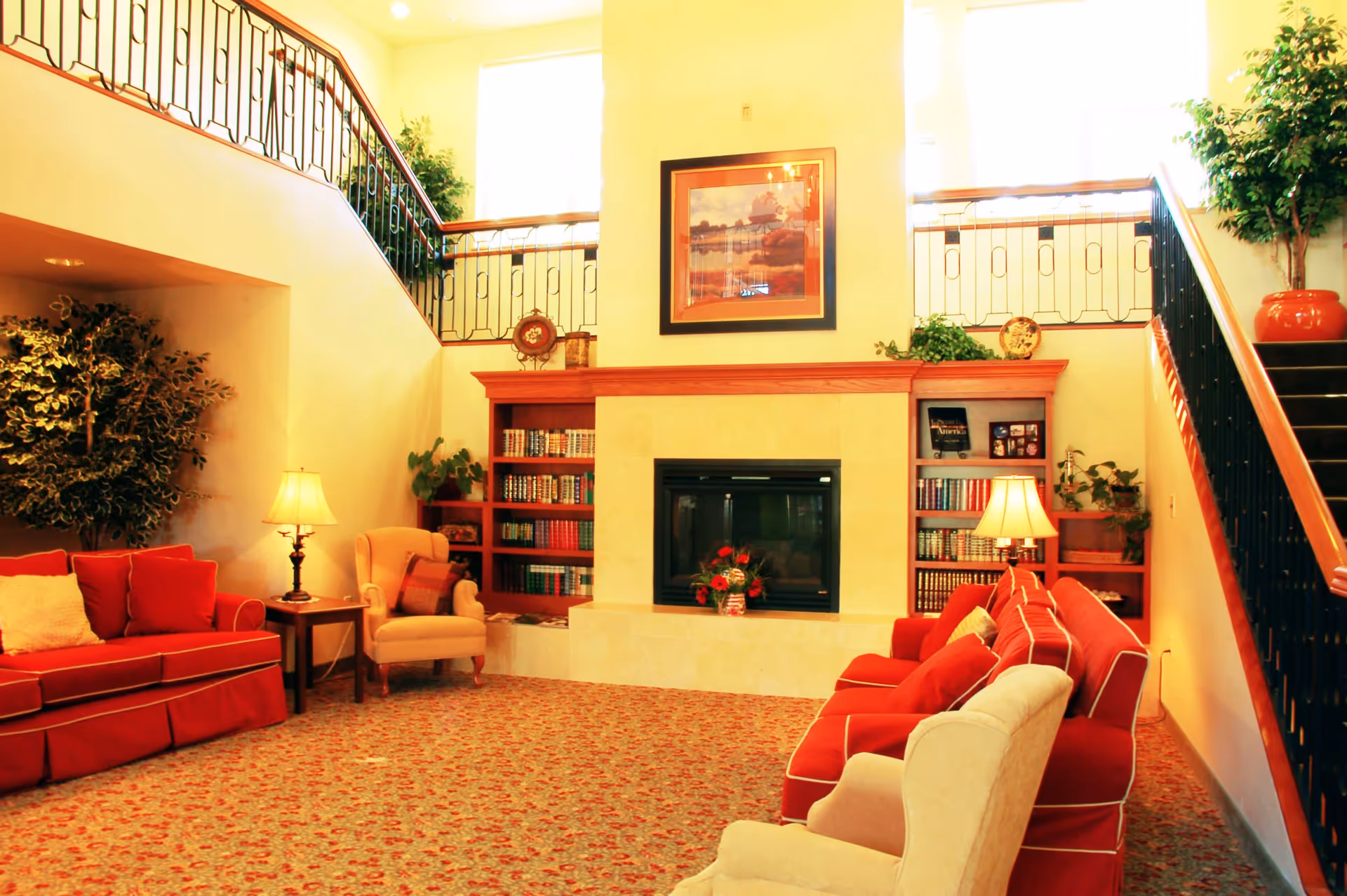 A cozy living room area with red sofas and beige armchairs arranged around a fireplace. The fireplace is flanked by built-in wooden bookshelves filled with books and decorative items. There are two table lamps on either side of the seating area, and large windows above let in natural light. A staircase with a black metal railing leads to an upper level, and there are potted plants adding greenery to the space.