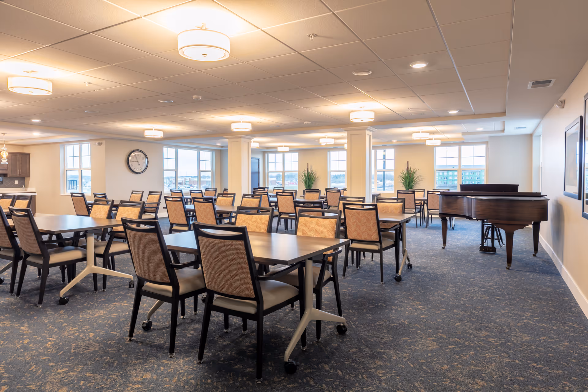 A spacious dining room with multiple tables and chairs arranged neatly. The room has large windows letting in natural light, ceiling lights, a wall clock, two tall green plants, and a grand piano in the corner.
