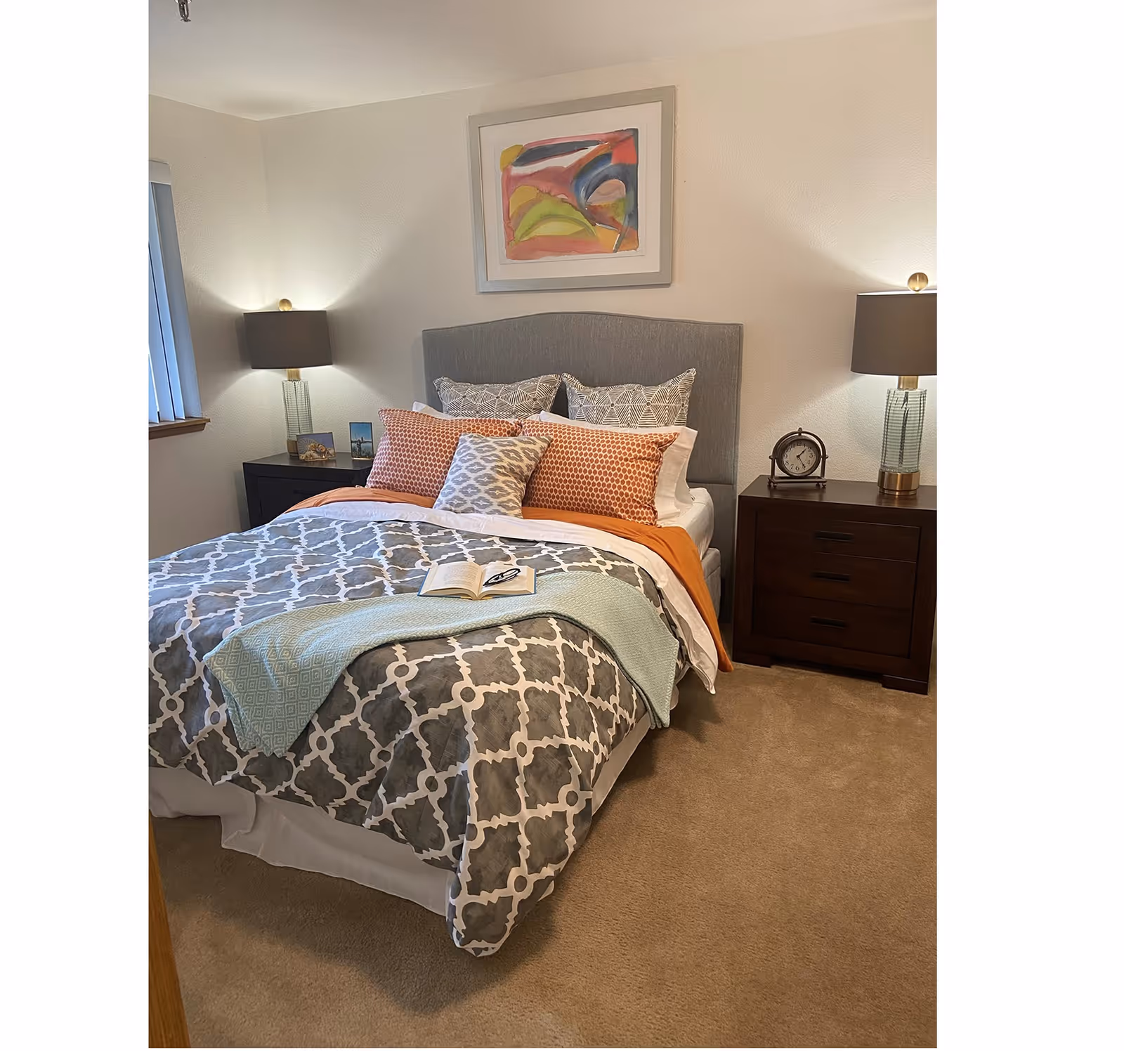 A neatly made bed in a bedroom with a gray patterned comforter, multiple decorative pillows in orange and gray tones, and a light blue throw blanket. There are two dark wooden nightstands on either side of the bed, each with a lamp. One nightstand also has a small clock and framed pictures. A colorful abstract painting hangs on the wall above the bed.