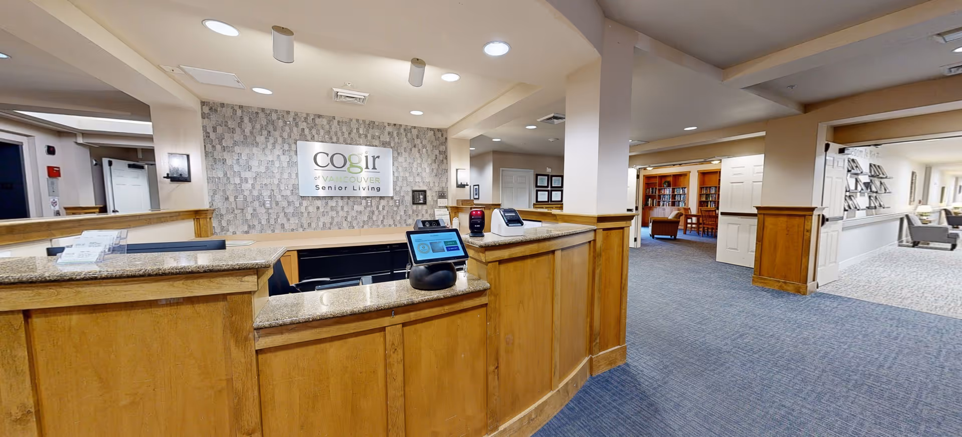 Reception area of a senior living facility with a wooden front desk and granite countertop. Behind the desk is a wall with a sign that reads 'cogir of Vancouver Senior Living'. The area is well-lit with recessed ceiling lights. To the right, there is a hallway leading to a room with bookshelves and seating, and further down another seating area is visible.