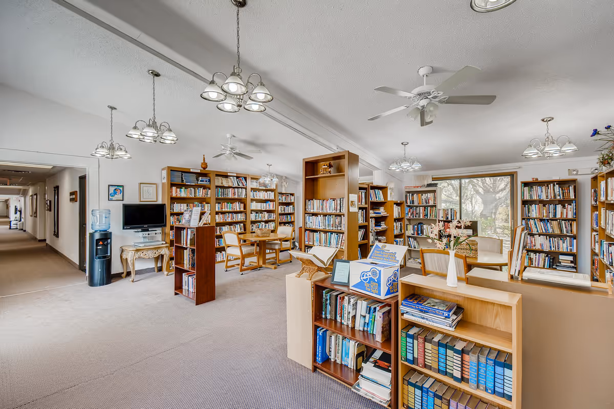 Bright senior living library with bookshelves, tables, chairs, and a hallway in the background.